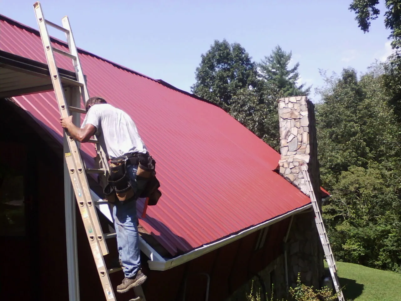 Roofer on a ladder beside a red metal roof and stone chimney — Soffit & Fascia Repair by Roofing Brothers in Keyes